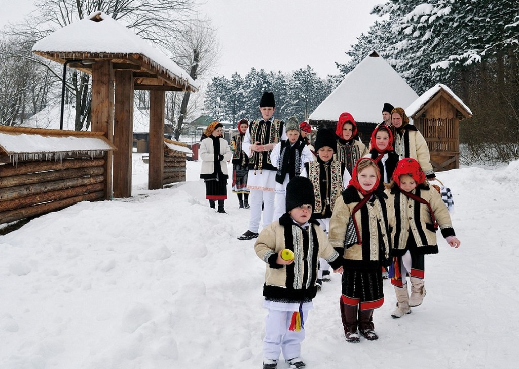 Imaginea surprinde un grup de copii și adulți îmbrăcați în port popular tradițional bucovinean, într-un cadru de iarnă autentic. Decorul este specific satelor tradiționale din Bucovina, cu case din lemn și zăpadă proaspăt căzută. Copiii poartă cojoace și cămăși cu broderii detaliate, fetele având baticuri viu colorate, iar băieții căciuli din blană. Adulții din spate, îmbrăcați în haine tradiționale, completează atmosfera autentică. Această imagine evocă frumusețea obiceiurilor bucovinene de iarnă, cum ar fi colindatul, și simbolizează legătura puternică a comunității cu tradițiile străvechi