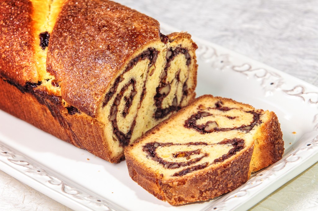 Cozonac tradițional românesc cu nucă și cacao, așezat pe o farfurie decorativă albă. Cozonacul este tăiat pentru a se vedea straturile spiralate de umplutură, având o crustă aurie și crocantă