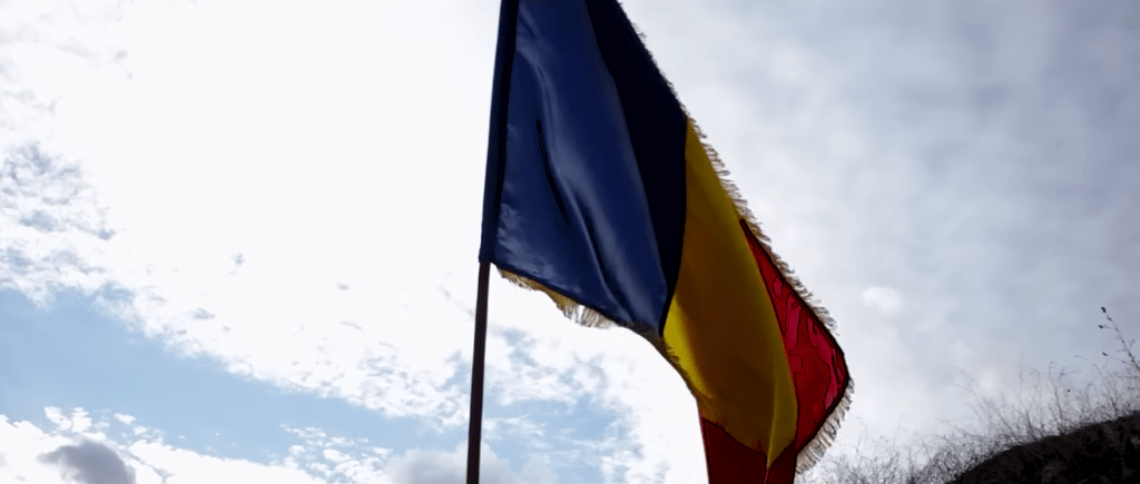 Tricolorul României fluturând în bătaia vântului, simbol al mândriei naționale și al valorilor autentice românești, sub cerul senin