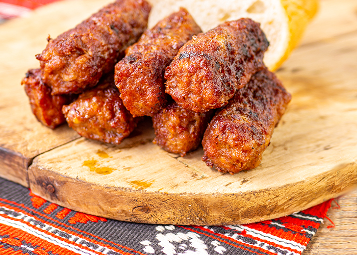 Porție de mici românești la grătar, serviți pe un tocător de lemn, alături de o felie de pâine, pe un ștergar tradițional cu motive populare